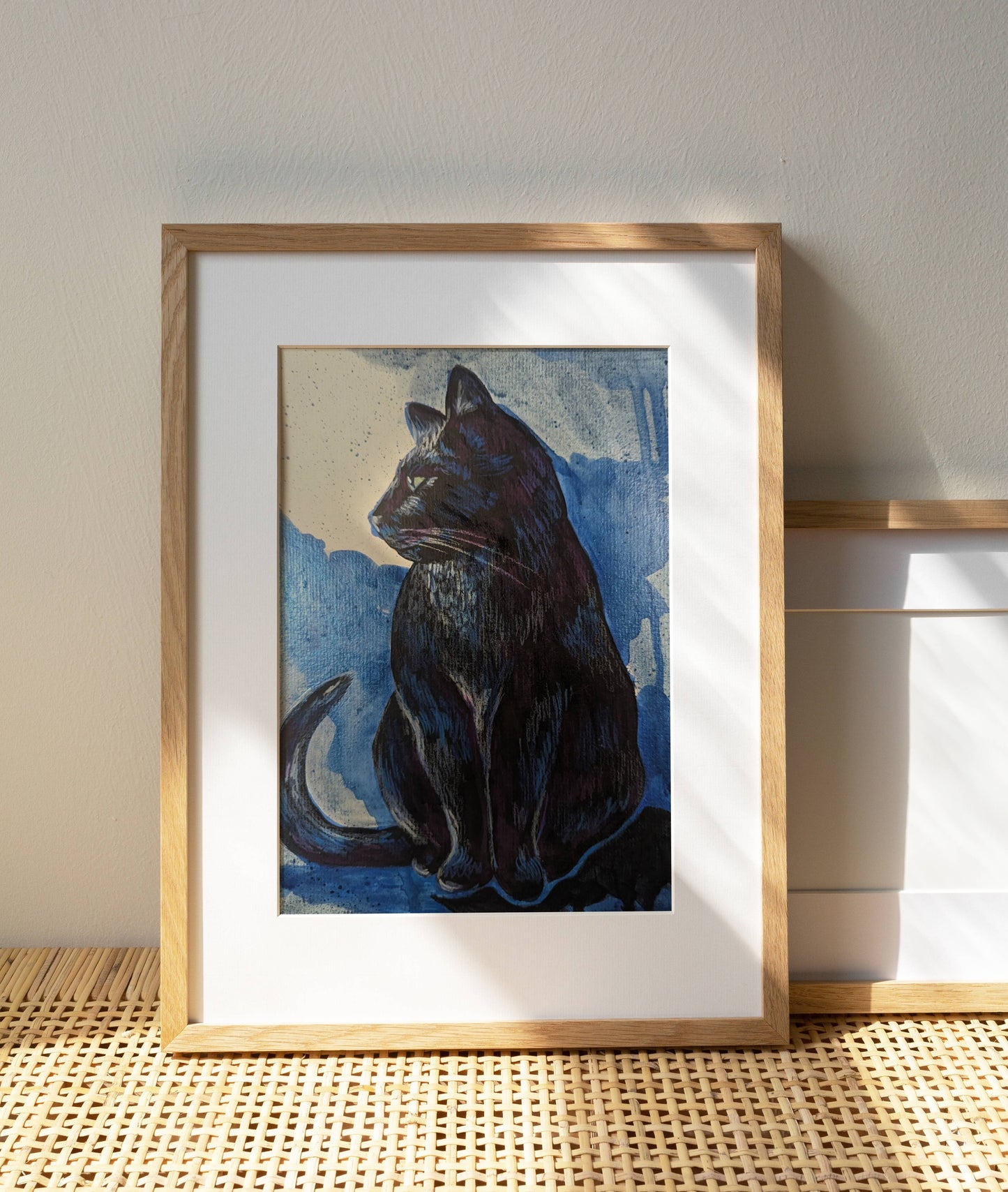 A framed black cat art print sitting on a woven rattan shelf with sunlight casting shadows on the wall