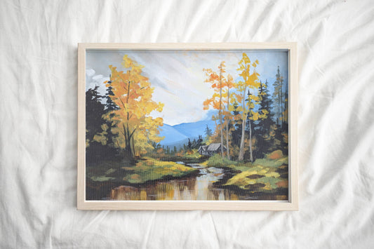 A framed art print of an autumn landscape with vibrant golden-yellow trees by a river with distant mountains in the background under a cloudy sky