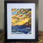 A framed sunset art print named Harmony leaning against a wall on a wooden surface